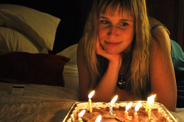 photo of Katrina Schaag with her chin propped on her palm and overlooking a cake with candles burning. The photo has a orange glow from the candlelight which casts half of Katrina Schaag's face in a warm glow and half in shadow